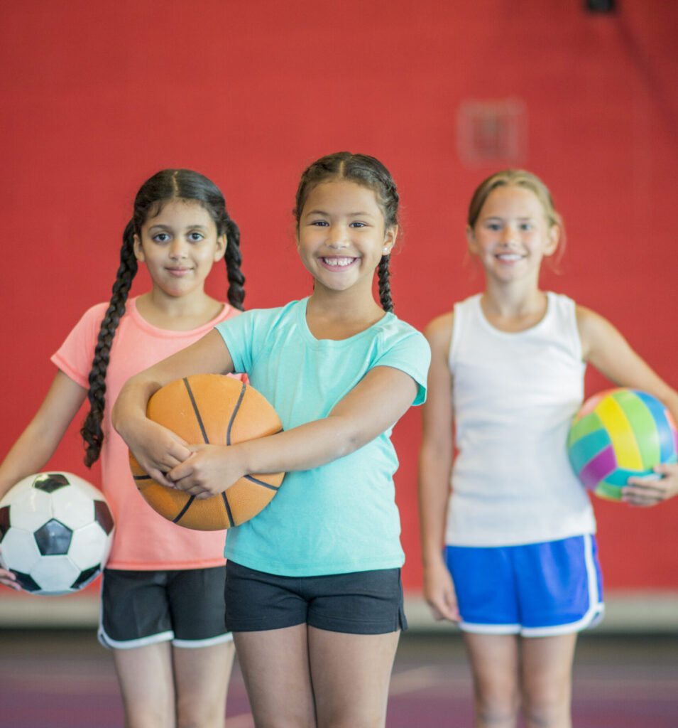 Photo of three girls 10 year old girls, one in orange top and black shorts holding a soccer ball, middle girl wearing a blue top and black shorts holding a basketball, and one girl wearing white singlet with blue shirts holding a coloured volleyball